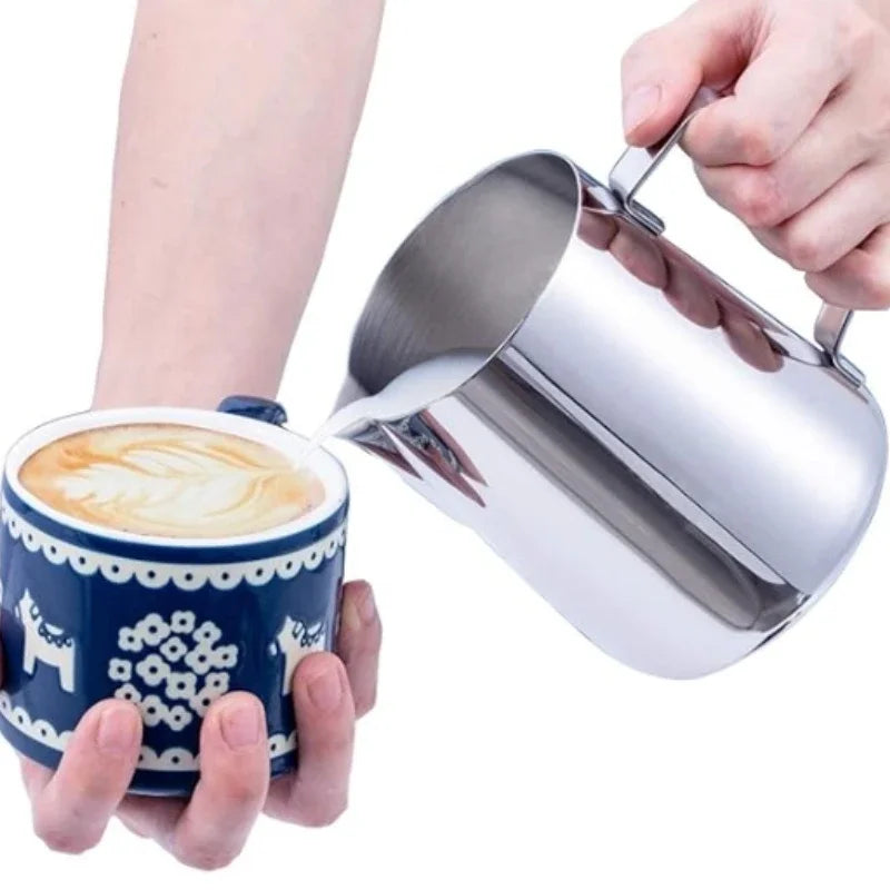 Hand holding a stainless steel milk frothing jug over a coffee cup for latte art preparation